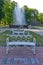 Elegant white bench with a curved back standing in a park near a green lawn in front of a beautiful fountain with clear
