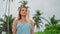 Elegant woman in blue dress strolls on sandy beach, admires serene tropical setting. Blonde explores coastline, reflects