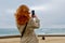 Elegant woman with red hair holding mobile phone and making photo of seaside on grey cloudy day