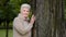 Elegant woman retirement age resting in park alone, standing near large tree, stroking it, touches bark tenderly