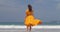Elegant young lady enjoying good day near ocean wearing long yellow dress, walking slowly on wet sand back view