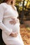 Elegant young pregnant woman in white knit dress and hat touching and stroking belly, relaxing in park on autumn warm