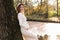 Elegant young pregnant woman in white knit dress and hat touching and stroking belly, relaxing in park by the lake on