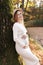 Elegant young pregnant woman in white knit dress and hat touching and stroking belly, relaxing in park by the lake on