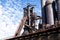 Elevated walkway alongside steel mill blast furnaces against a blue sky with clouds