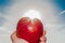 Elongated hand of a man holding a red apple against a blue sky and sun