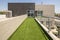 Elongated terrace in the shape of an L covered with artificial grass overlooking a summer pool and in front of the facade of the