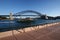 Embankment waterfront promenade with ferry cruising by iconic Sydney Harbor Bridge. Coastal cityscape with historic landmark.