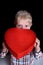 Embarrassed blond boy holding a red box in the shape of a heart on black background. Close up