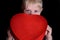 Embarrassed blond boy holding a red box in the shape of a heart on black background. Close up