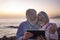 Embraced attractive senior couple white-haired looking at digital tablet in the dusk sitting on the beach with horizon over water