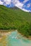 Emerald green water of Socca River flowing over rocky riverbed with green mountain background in Western Slovenia