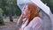 Emotional fat ginger girl with glasses is standing under rain in park, talking on phone, holding umbrella, communication