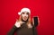 Emotional girl in santa hat shows finger on smartphone with screen, looks at camera,smiles on red background. Astonished lady