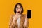 Emotional lady showing smartphone with blank screen and covering her mouth in shock on yellow background, mockup