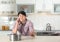 Emotional man calling plumber near table with saucepan under leaking water from ceiling