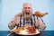 Emotional obese man holds sausage and smoked chicken leg sitting at table