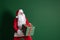 Emotional Santa Claus sneaking while holding big green gift box in hands on green studio background