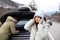 Emotional young woman talking on phone while stressed man repairing broken car in winter
