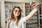 Emotive fashionable urban woman in optician store standing over stands with glasses while taking selfie in stylish