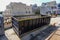 An empty black dumpster is seen at a construction site near a backhoe