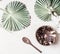 Empty coconut bowl with spoon on white desk background with tropical leaves, top view. Copy space for your design or product