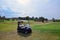 An empty golf cart stands on the lawn of a private golf club. Behind him is a large clearing and a blue sky. Active summer luxury