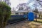 Empty green dumpster with a blue portable potty are seen in a driveway in front of a small house in a residential neighborhood.