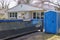 Empty green dumpster with a blue portable potty are seen in a driveway in front of a small house in a residential neighborhood.