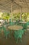 Empty restaurant under a white canopy among green palm trees with marble tables and green wrought-iron chairs