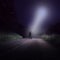 An empty road at night with a lone figure looking up at bright UFO with a white beam of light coming down