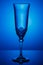 Empty transparent champagne glass on a thin high leg in dark blue backlight with reflection on a glossy surface