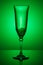 Empty transparent champagne glass on a thin high leg with reflection on a dark glossy surface in green backlight