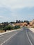 Empty two lane road with pavements and lampposts curving up a hill into the distance with surrounding trees and blue sunlit sky