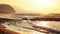 Empty wild beach lit by golden sunset. Small waves over rock, hills in hazy background