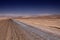 Endless dirt road to infinity of salt flat plateau contrasting with blue cloudless sky.