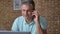 Energetic adult caucasian male is talking over phone and using his computer while sitting in office, brown brick