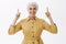 Energized active and happy old woman with white hair in stylish yellow coat raising index fingers pointing up and