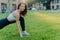 Energized woman in top and leggings, makes stretching excerises at green lawn during sunny day outdoor, warms up before cardio
