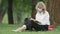 Engrossed middle aged blond woman in eyeglasses and coronavirus face mask reading book sitting on summer meadow in park