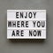 `Enjoy where you are now` words on lightbox over concrete background, top view. Flat lay, from above, overhead
