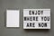 `Enjoy where you are now` words on modern board, notepad over concrete background, top view. Flat lay, from above. Close-up