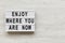 `Enjoy where you are now` words on modern board over white wooden background, overhead. Copy space