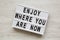 `Enjoy where you are now` words on modern board over white wooden surface, overhead view. Flat lay, from above