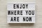`Enjoy where you are now` words on modern board over white wooden surface, top view. Close-up