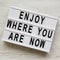 `Enjoy where you are now` words on modern board over white wooden surface, top view. Flat lay, from above