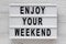 `Enjoy your weekend` words on a lightbox on a white wooden background, top view. Overhead, from above, flat lay