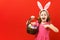 Enthusiastic child in the ears of a bunny on a red background. Little girl shows a finger on a basket with Easter eggs