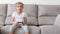 Enthusiastic kid in casual outfit playing with joystick while sitting on sofa in living-room.