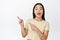 Enthusiastic korean girl pointing fingers left, looking amazed at camera, standing in yellow t-shirt over white